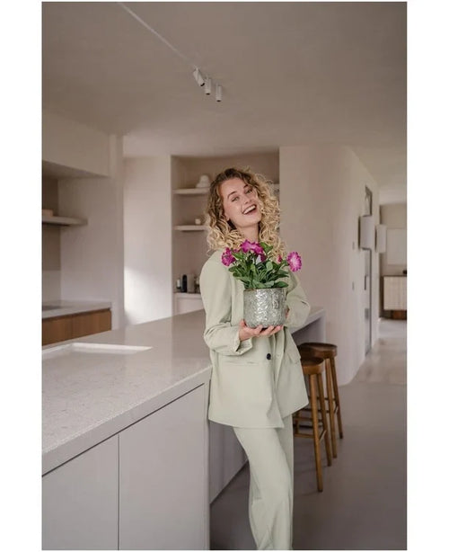 En leende kvinna med lockigt hår, iklädd en ljus kostym, står i ett modernt, minimalistiskt kök och håller i en Artificiell Blomma Petunia UV 25 cm mörkrosa med levande rosa blommor.
