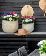 Två stora keramikkrukor med en konstgjord blomma Petunia UV 25 cm mörkrosa, med levande rosa blommor, står på ett bord mot en mörk trävägg. Det står en stråhatt på bordet och torkade palmblad hänger på väggen ovanför blommorna.