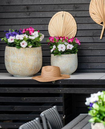 Två stora keramikkrukor med en konstgjord blomma Petunia UV 25 cm mörkrosa, med levande rosa blommor, står på ett bord mot en mörk trävägg. Det står en stråhatt på bordet och torkade palmblad hänger på väggen ovanför blommorna.