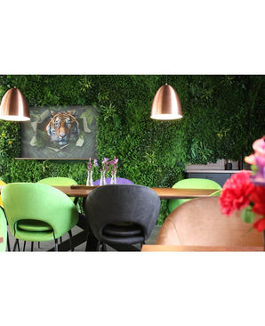 Een moderne eethoek met groene en perzikkleurige stoelen, een tafel versierd met vazen met paarse bloemen, koperen hanglampen, een weelderige plantenwand gemaakt met het Kunsthaag Crush UV-paneel van 100 x 100 cm, en op de achtergrond een ingelijste afbeelding van een tijger.