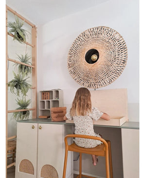 Ett litet barn med axellångt hår sitter vid ett skrivbord i ett ljust rum, med ryggen vänd mot betraktaren. Bakom henne finns en vävd vägglampa, några skåp och den konstgjorda hängande växten 'Frosted Fern on ball 76 cm', inbäddad bland växterna på en trästege, bredvid små hyllor med snyggt arrangerade föremål.
