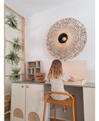 Ett litet barn med axellångt hår sitter vid ett skrivbord i ett ljust rum, med ryggen vänd mot betraktaren. Bakom henne finns en vävd vägglampa, några skåp och den konstgjorda hängande växten 'Frosted Fern on ball 76 cm', inbäddad bland växterna på en trästege, bredvid små hyllor med snyggt arrangerade föremål.