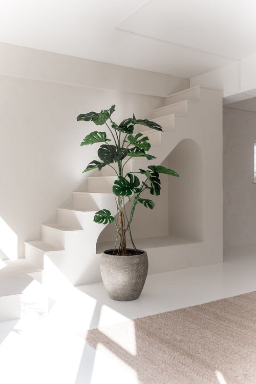 Den konstgjorda växten Monstera 210 cm, med breda gröna blad, står på ett vitt golv i ett ljust, minimalistiskt rum med vita väggar, en trappa och naturligt solljus.