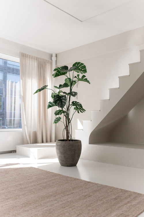 Den konstgjorda växten Monstera 210 cm står i en kruka på ett vitt golv bredvid en modern trappa i ett ljust, minimalistiskt rum med naturligt ljus som kommer in genom beiga gardiner.