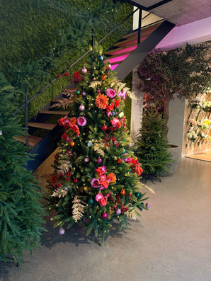 Den konstgjorda julgranen "Weideman" PE/PVC - Smart LED är placerad inomhus nära en trappa, dekorerad med rosa och röda blommor, bladguld och rosa prydnader, omgiven av grönska på ett polerat betonggolv för en snygg juldekoration.
