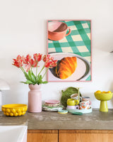 En bänkskiva med alla möjliga färgglada serviser och en konstgjord blomma French Parakit Tulpan på 76 cm i rött/gult i en rosa vas. Ovanför den hänger en inramad bild av en croissant och en tekopp på en grönrutig duk.