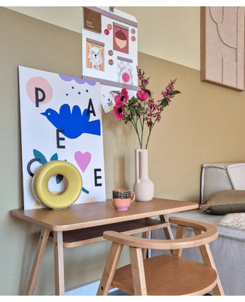 Ett litet träskrivbord med stol är dekorerat med Artificiell Blomma Kattmynta 75 cm rosa, en mugg, en gul ringskulptur och färgglada konsttryck på väggen ovanför. En bänk är delvis synlig till höger.