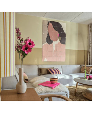 Een moderne woonkamer met een modulaire bank, een roze bloemstuk van Kunstbloem Catnip (75 cm) in een vaas, een donzige plaid en een abstract wandschilderij van een vrouw. Het interieur wordt gekenmerkt door zachte kleuren en accenten met patronen.
