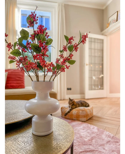 På ett bord står en vit vas med rosa Kunsttak-bär (64 cm) och gröna blad. I bakgrunden ligger en katt på en rosa kudde i ett ljust vardagsrum med beiga väggar och en glasdörr.