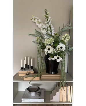 På en hylla mot en ljus vägg står en svart vas med Konstblomman Amaranthus 124 cm grön, bredvid fem vita ljus, beiga böcker och prydnadsföremål i neutrala toner.