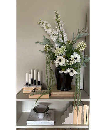På en hylla mot en ljus vägg står en svart vas med Konstblomman Amaranthus 124 cm grön, bredvid fem vita ljus, beiga böcker och prydnadsföremål i neutrala toner.