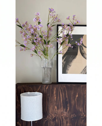 I en transparent vas placeras blommorna i Kunsttak Daphne Spray 108 cm lila på en mörk träyta bredvid ett modernt, abstrakt konstverk i en ram, med en vit strukturerad lampskärm under.