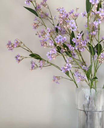 Raffinerad artificiell gren Daphne-gren 108 cm i lila, med gröna blad, arrangerad i en klar, räfflad glasvas och placerad mot en enkel, ljus bakgrund.