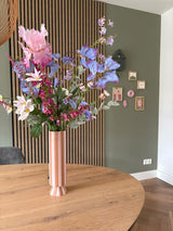 Een hoge roze en wit gestreepte vaas met het zijden kunstbloemen boeket Amethyst Romance staat op een ronde houten tafel in een moderne kamer met groene muren, houten lambrisering en kleine ingelijste kunst.