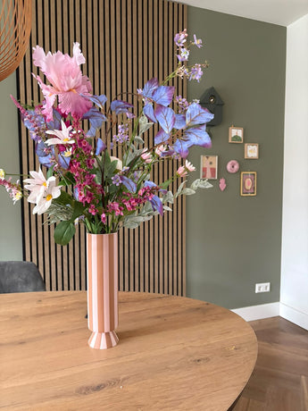 Een hoge roze en wit gestreepte vaas met het zijden kunstbloemen boeket Amethyst Romance staat op een ronde houten tafel in een moderne kamer met groene muren, houten lambrisering en kleine ingelijste kunst.