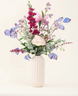 En vas med vit textur visar upp "Silk Artificial Flowers Bouquet Amethyst Romance" med rosa pioner, lila och blå blommor, vinröda blommor och grönt blad mot en ljus, neutral bakgrund.