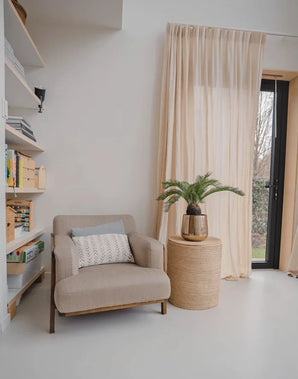 En mysig läshörna med en beige fåtölj, ett runt sidobord i flätat med konstgjorda Baby Cycas Palm 60 cm växt på, bokhyllor och nätgardiner som hänger över en glasdörr som vetter mot trädgården.