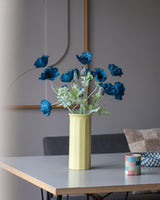 En 73 cm konstgjord blomvallmo i mörkblått står i en gul vas på ett bord, ackompanjerad av en liten färgglad randig kopp, i ett modernt, mjukt upplyst rum med minimalistisk inredning och neutrala toner.