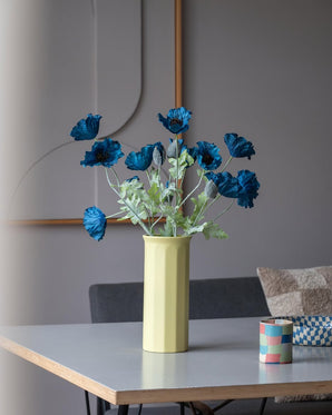 En 73 cm konstgjord blomvallmo i mörkblått står i en gul vas på ett bord, ackompanjerad av en liten färgglad randig kopp, i ett modernt, mjukt upplyst rum med minimalistisk inredning och neutrala toner.