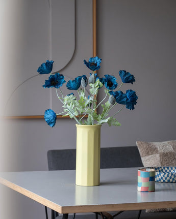 En 73 cm konstgjord blomvallmo i mörkblått står i en gul vas på ett bord, ackompanjerad av en liten färgglad randig kopp, i ett modernt, mjukt upplyst rum med minimalistisk inredning och neutrala toner.