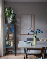 Den moderna matsalen har ett grått vitrinskåp i vilket porslin och växter visas upp, minimalistiska väggdekorationer och ett bord på vilket Konstgjorda blomstervallmo 73 cm mörkblå står i en gul vas. Utrymmet kompletteras av en eklektisk blandning av stolar, allt mot en bakgrund av grå väggar och ett trägolv.