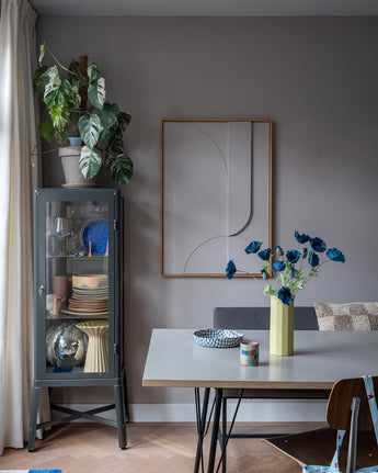 Den moderna matsalen har ett grått vitrinskåp i vilket porslin och växter visas upp, minimalistiska väggdekorationer och ett bord på vilket Konstgjorda blomstervallmo 73 cm mörkblå står i en gul vas. Utrymmet kompletteras av en eklektisk blandning av stolar, allt mot en bakgrund av grå väggar och ett trägolv.