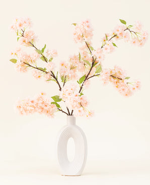 Een witte keramieken vaas met een brede ovale opening toont verschillende groenbladige Zijden kunstbloemen boeket Kersenbloesem lichtroze takken tegen een eenvoudige lichte achtergrond.