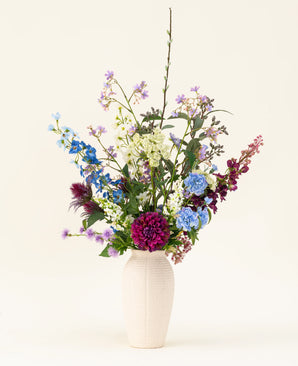 En vit vas med den konstgjorda sidenblommabuketten Purple Rain visar levande lila och vita konstgjorda blommor och gröna blad, ett exempel på holländskt hantverk mot en ljus bakgrund.