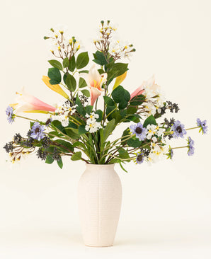 In een witte vaas staat het Zijden kunstbloemen boeket Blush & Bloom - met roze lelies, witte en paarse bloemen en weelderige groene bladeren - tegen een lichte achtergrond.