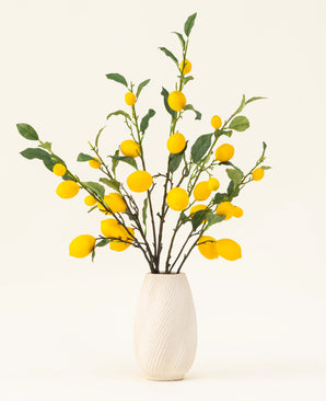 Een vaas met witte structuur toont het Zijden kunstbloemen boeket Lemons, met groene bladeren en levendige gele citroenen tegen een lichte achtergrond.