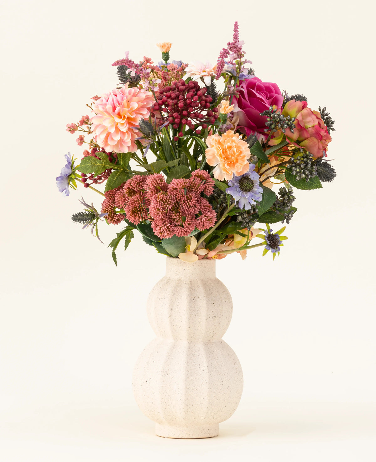 I en vit, böjd keramikvas finns den konstgjorda blombuketten Pretty Pastel siden - en färgstark blandning av hållbara blommor som rosa rosor, dahlior, lila och orange blommor och grönska - vackert arrangerad mot en ljus bakgrund.