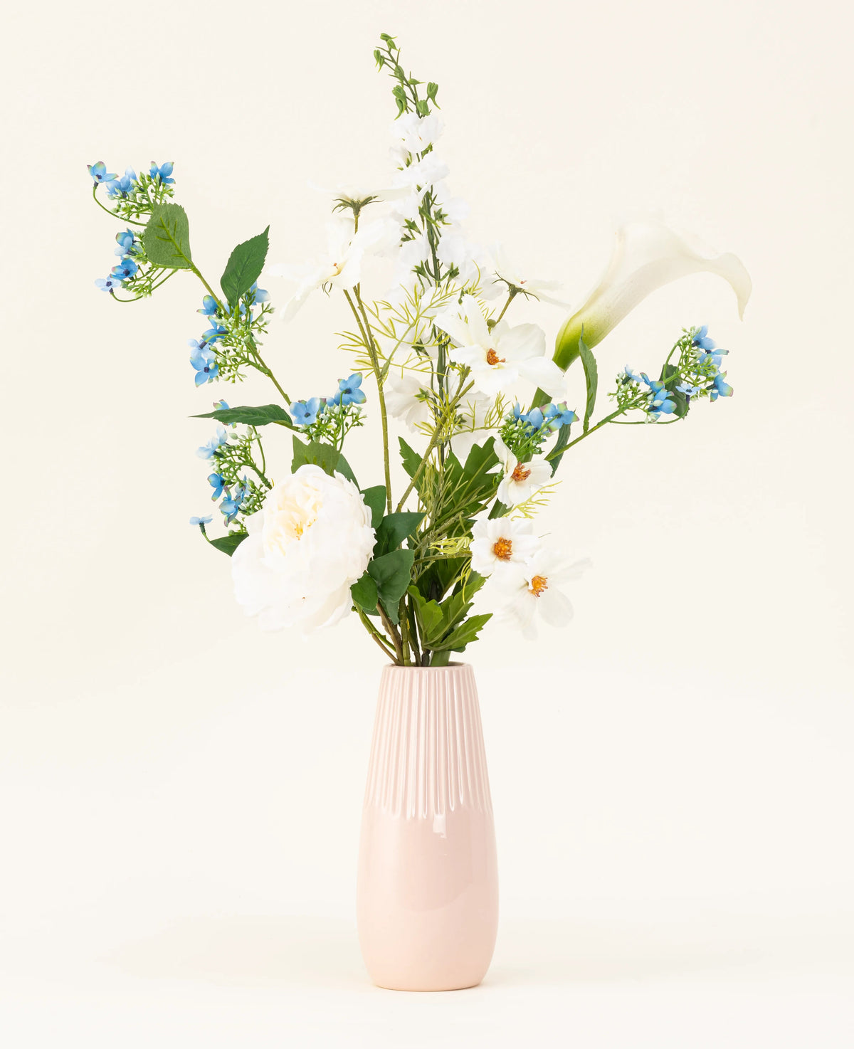 Den ljusrosa vasen innehåller konstgjorda blombuketten Whispers of White siden med vita blommor, blå blommor och grönt blad mot en enkel, ljus bakgrund.