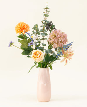 Het Zijden kunstbloemen boeket Earth & Petal is een pastelroze vaas met levensechte zijden bloemen in oranje, roze en geel, plus groen en paarse accenten, afgebeeld tegen een lichte achtergrond.