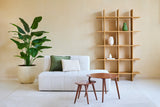 Minimalistiskt vardagsrum med en vit soffa, två kuddar, häckbord i trä och en stor konstgjord växt Strelitzia Real Touch Deluxe 180 cm för en tropisk atmosfär. En modern öppen bokhylla i trä med vaser står mot den beiga väggen.