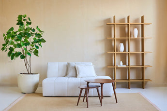 Minimalistische woonkamer met een witte bank, twee geneste houten salontafels, beige muren, een open houten plank met witte vazen en een Kunstplant Ficus 180 cm die groen toevoegt. Een beige vloerkleed bedekt de vloer.
