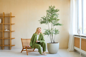 En kvinna i grön kostym ler när hon sitter på en trästol bredvid växten Artificiell Ruscus 180 cm i ett ljust, modernt rum med beiga väggar, stora fönster och minimalistisk inredning.