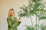 Een glimlachende vrouw in een groene blazer staat binnen en raakt voorzichtig de bladeren aan van een Kunstplant Ruscus 150 cm met dunne takken en weelderig groen gebladerte, dat afsteekt tegen een effen, lichtgekleurde achtergrond.