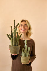 En leende kvinna i en brun klänning håller två Artificiella Cactus Cereus 35 cm krukväxter, en stor och en liten, mot en enkel beige bakgrund.
