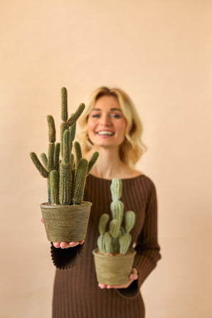 En leende kvinna i en brun klänning håller två Artificiella Cactus Cereus 35 cm krukväxter, en stor och en liten, mot en enkel beige bakgrund.