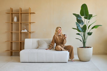En kvinna i en brun outfit sitter på en vit bank, glimtar med en kopia i handen. Bredvid henne finns den konstgjorda växten Strelitzia 180 cm verklig touch, som för frodig grönska in i det moderna, beige-tonade vardagsrummet med minimal inredning.
