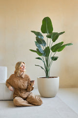 En kvinna i brun jumpsuit sitter leende på golvet med en kaffekopp bredvid sig, bredvid den konstgjorda växten Strelitzia 180 cm äkta touch i ett minimalistiskt rum med beiga väggar.