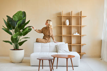 En kvinna i brun jumpsuit sitter på baksidan av en vit soffa och rör vid bladet på en Artificiell Strelitzia Real Touch Deluxe 180 cm växt i ett minimalistiskt vardagsrum med träbord och hyllor.