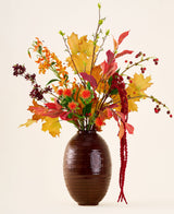 I en brun vas finns 'Rustic Glow'-buketten av konstgjorda sidenblommor, med gula, orange och röda blad, röda bär, orange blommor och långa, hängande röda amaranthus mot en neutral bakgrund.