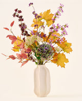 'Wild Amethyst'-buketten med konstgjorda sidenblommor, med höstlöv, lila och vita blommor och gröna hortensior, är i en vit keramikvas mot en enkel, ljus bakgrund.