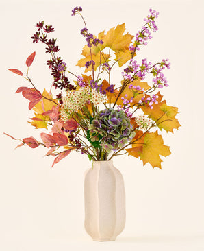 'Wild Amethyst'-buketten med konstgjorda sidenblommor, med höstlöv, lila och vita blommor och gröna hortensior, är i en vit keramikvas mot en enkel, ljus bakgrund.