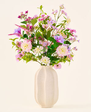 En vit keramikvas rymmer en färgstark bukett med rosa, lila och vita blommor från kollektionen 'Silk artificiell blombukett Royal Bloom', accentuerad med gröna blad och presenterad mot en enkel, ljus bakgrund.
