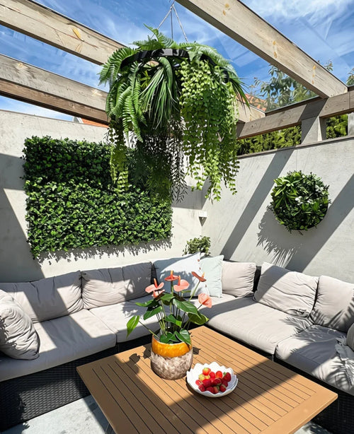 En solig terrass med beiga sittmöbler med kuddar och ett träbord, dekorerad med en ananasvas med rosa blommor och en skål med jordgubbar. Gröna väggväxter ger en frodig inredning, kompletterad med den konstgjorda hängväxtormbunken UV 60 cm i svart ring som en slående hängande accent.