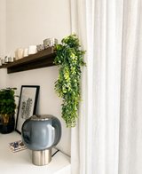 Een moderne woonruimte met een grijze glazen lamp, ingelijste kunst, een potplant en een houten plank met kopjes en de Kunst-hangplant ‘Pannenkoek’ (61 cm), waarvan de ranken naast witte gordijnen hangen.