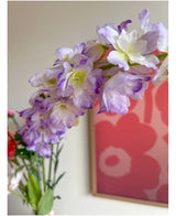 Närbild av den konstgjorda blomman Delphinium 105 cm lila, arrangerad i en vas, med ett rosa och rött blomkonstverk på väggen i bakgrunden.