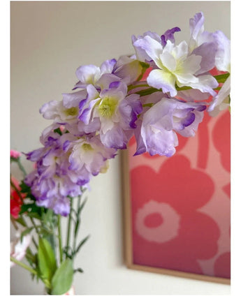 Närbild av den konstgjorda blomman Delphinium 105 cm lila, arrangerad i en vas, med ett rosa och rött blomkonstverk på väggen i bakgrunden.